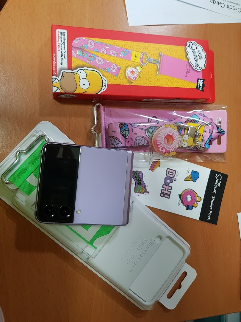 A purple Samsung Galaxy Z Flip phone, a silicone cover, and Simpsons-themed accessories are displayed.