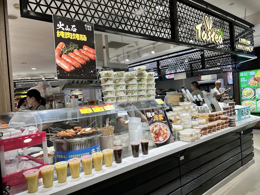 A vibrant food counter named "Tasted" offering grilled sausages, pre-packaged meals, and various beverages.
