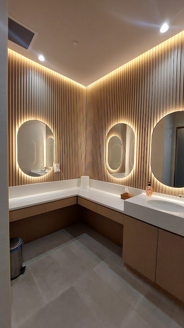 A modern, well-lit washroom interior with multiple sinks, illuminated mirrors, and wooden slat walls.
