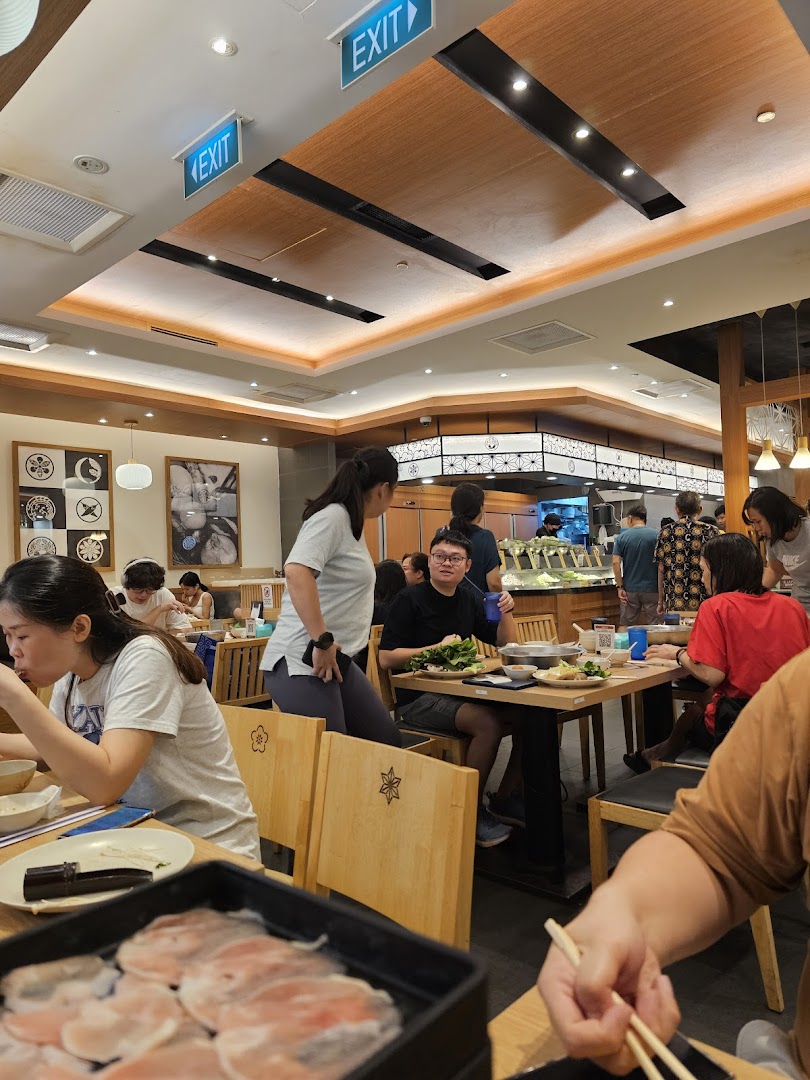 Diners enjoying shabu-shabu in a bustling restaurant interior with a buffet line visible.