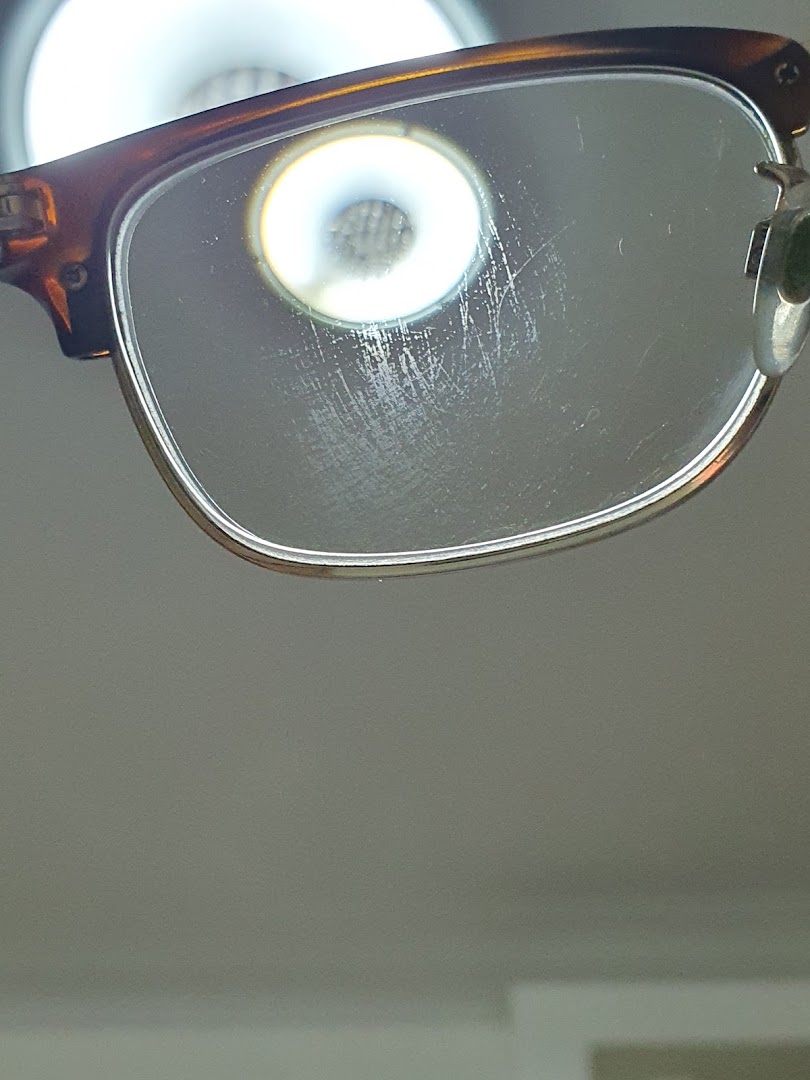 A close-up of an eyeglasses lens with visible scratches and a bright light reflection.