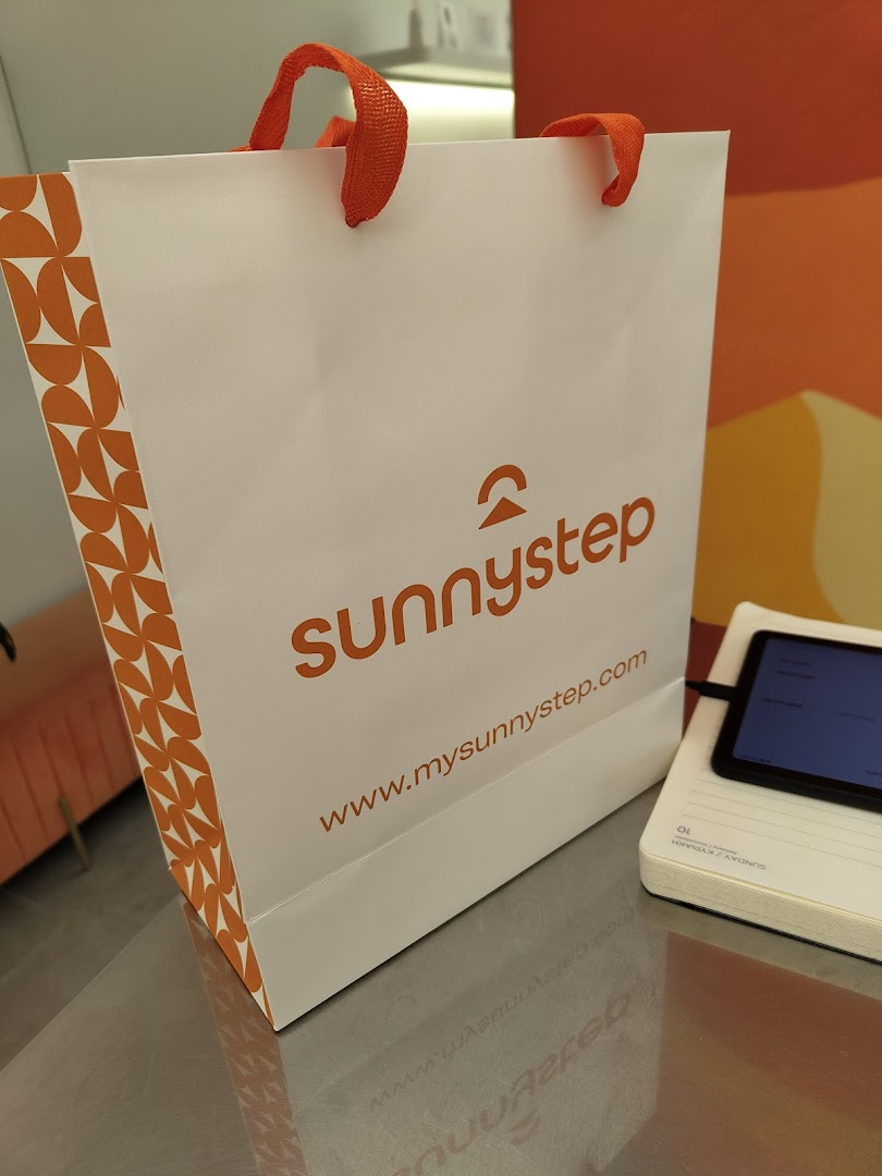 A white Sunnystep shopping bag with orange logo and handles, next to a tablet.