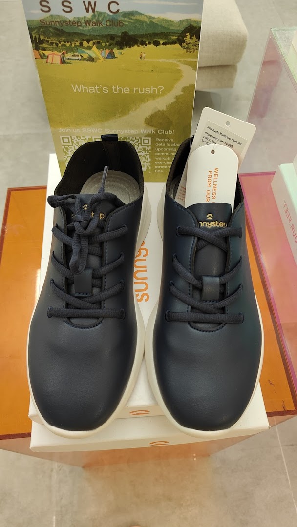 A pair of dark blue Sunnystep Balance Runner shoes displayed with a Sunnystep Walk Club promotional stand.