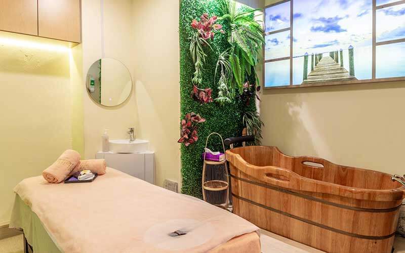 A serene spa room featuring a massage bed, wooden bath tub, green wall, and scenic window art.