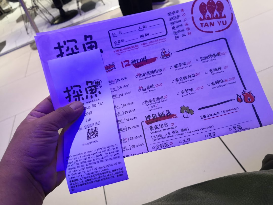 A hand holding a TANYU menu and queue ticket with various fish flavors and side dishes.