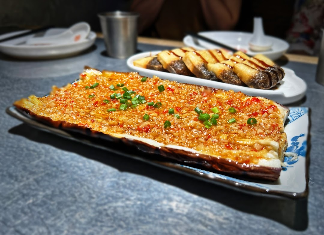 A close-up of grilled eggplant with garlic and chili, alongside another dish on a table.