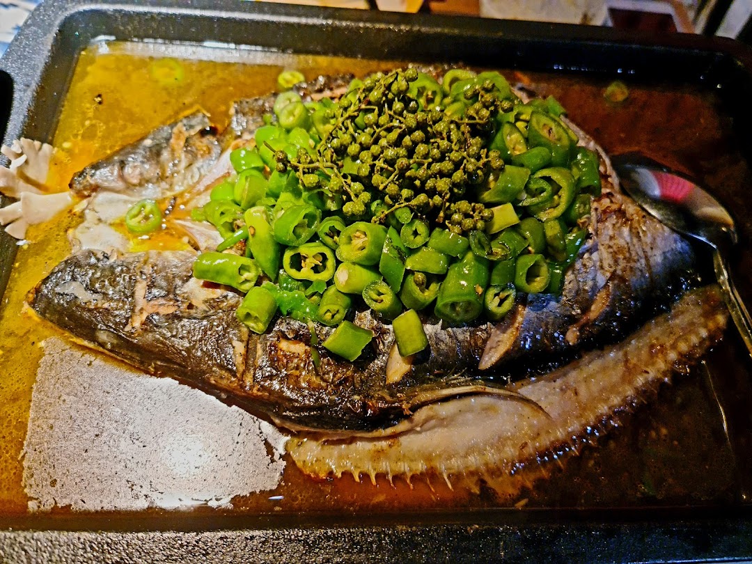 A close-up of a whole roasted fish dish, generously topped with green chili peppers and Sichuan peppercorns, served in a hot pan.