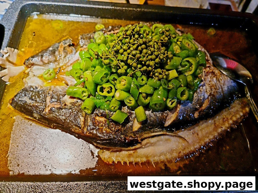 A close-up of a whole roasted fish dish, generously topped with green chili peppers and Sichuan peppercorns, served in a hot pan.