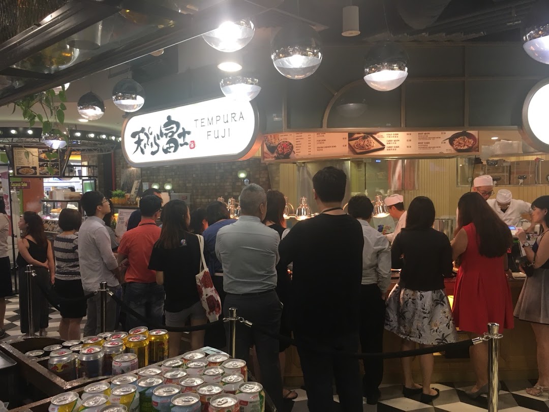 Customers queue at Tempura Fuji, watching chefs prepare fresh Japanese dishes in a bustling food court setting.
