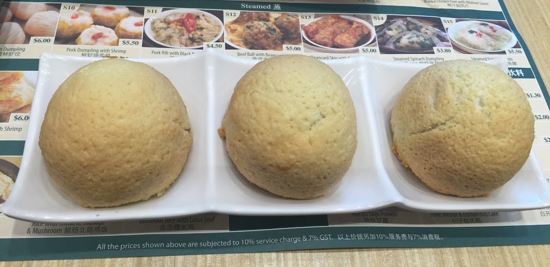 Three baked buns on a white plate with a dim sum menu board in the background.