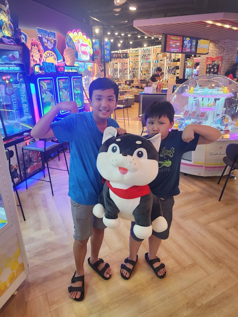 Two boys pose with a large plush toy inside a vibrant arcade with various game machines.
