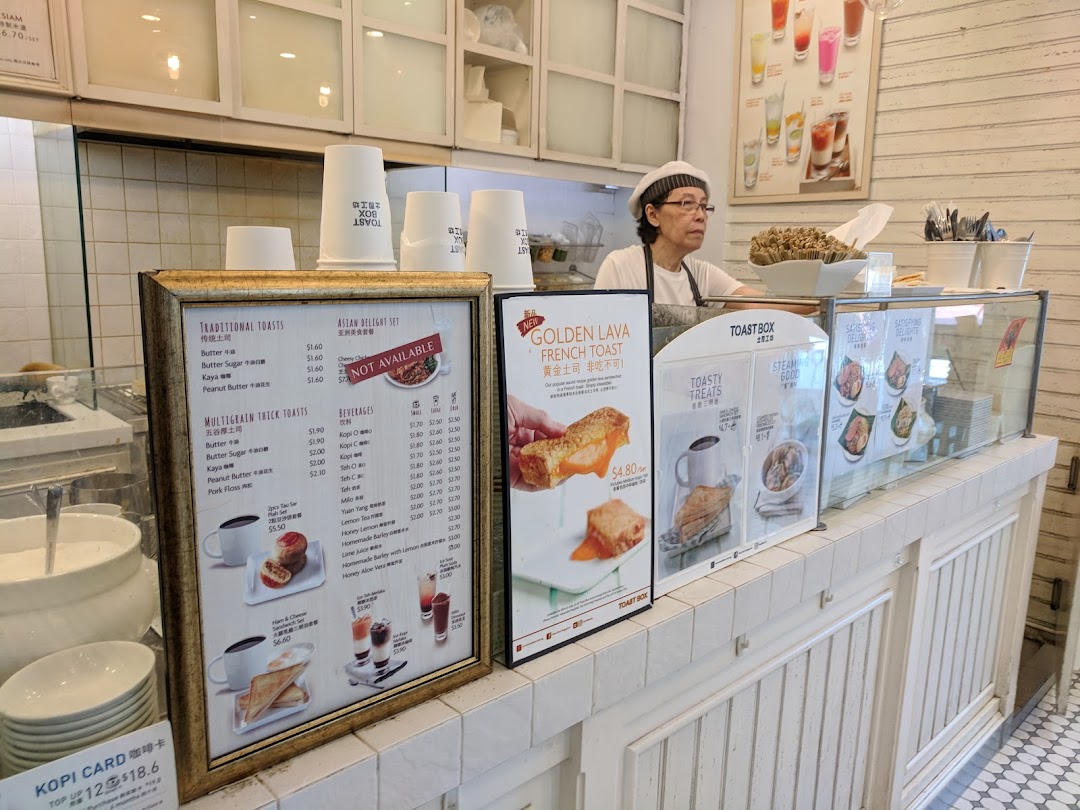 A staff member behind the counter at Toast Box, featuring prominent menu displays and food items.