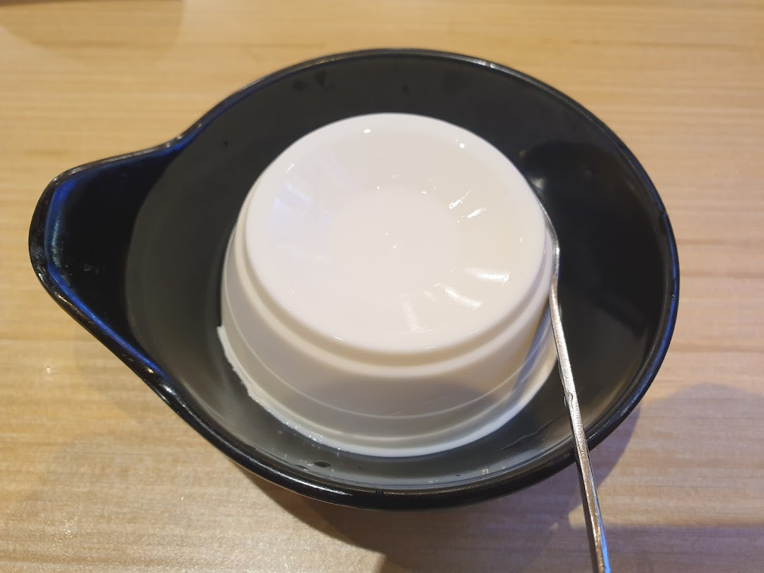 A smooth white dessert, possibly tofu pudding or panna cotta, served in a black bowl with a spoon.