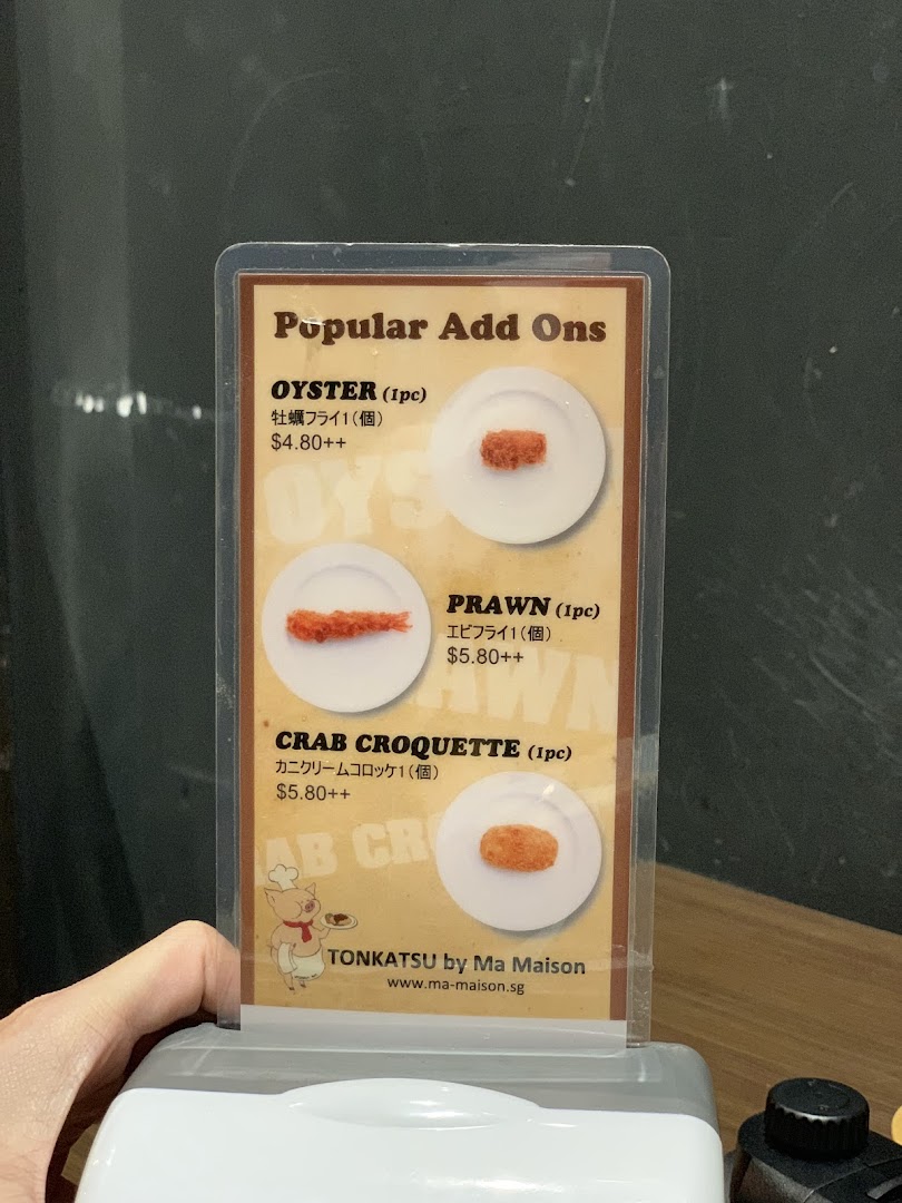 A table-top display showcasing popular add-on items and their prices from Tonkatsu by Ma Maison.