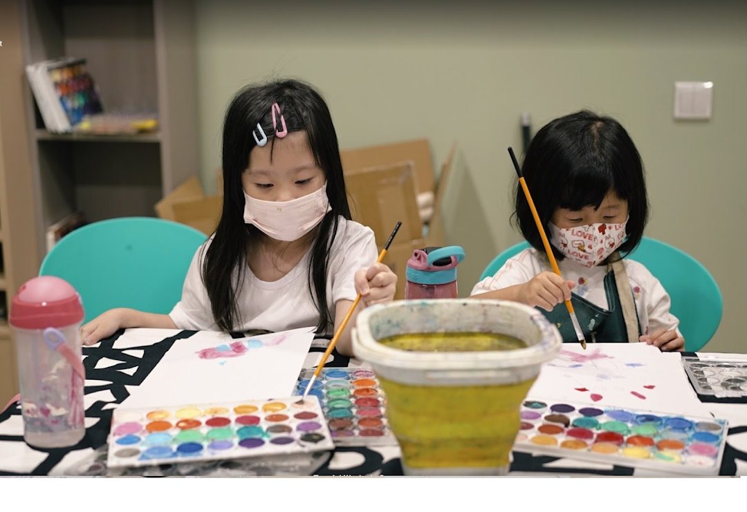 Two young girls wearing masks are engaged in watercolor painting at a table.