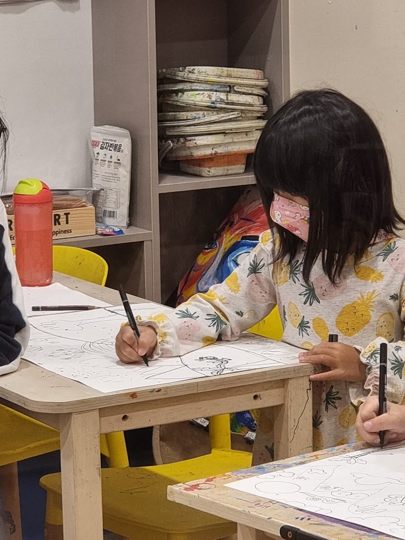A young child wearing a mask is focused on drawing at a table in an art studio, surrounded by art supplies.