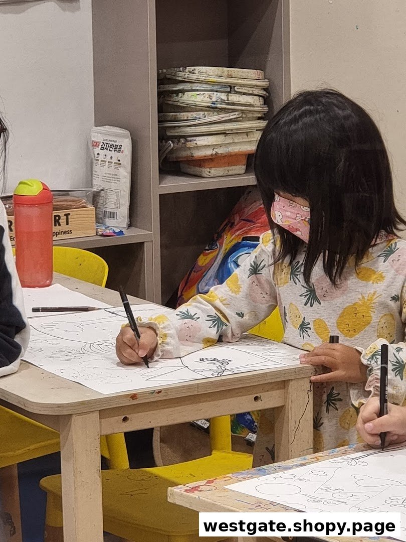 A young child wearing a mask is focused on drawing at a table in an art studio, surrounded by art supplies.