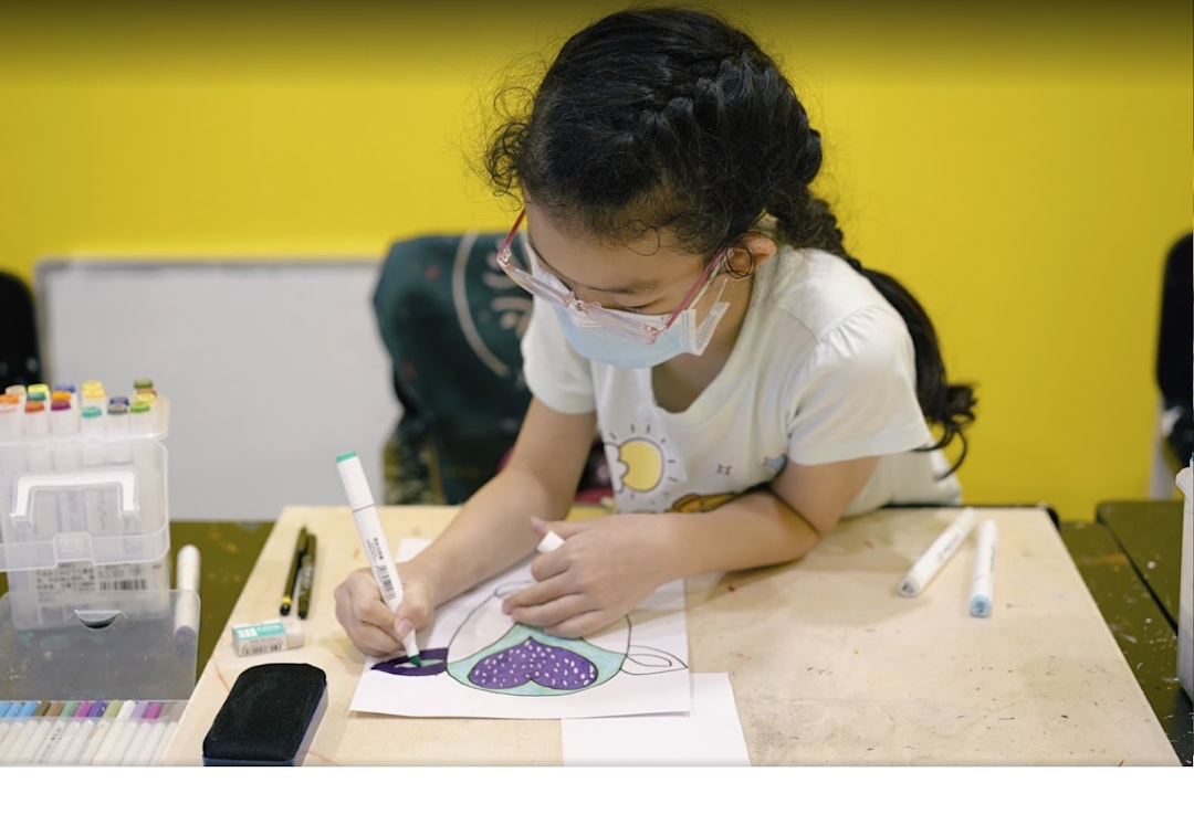A young student wearing a mask and glasses focuses on coloring an artwork with markers at a desk.