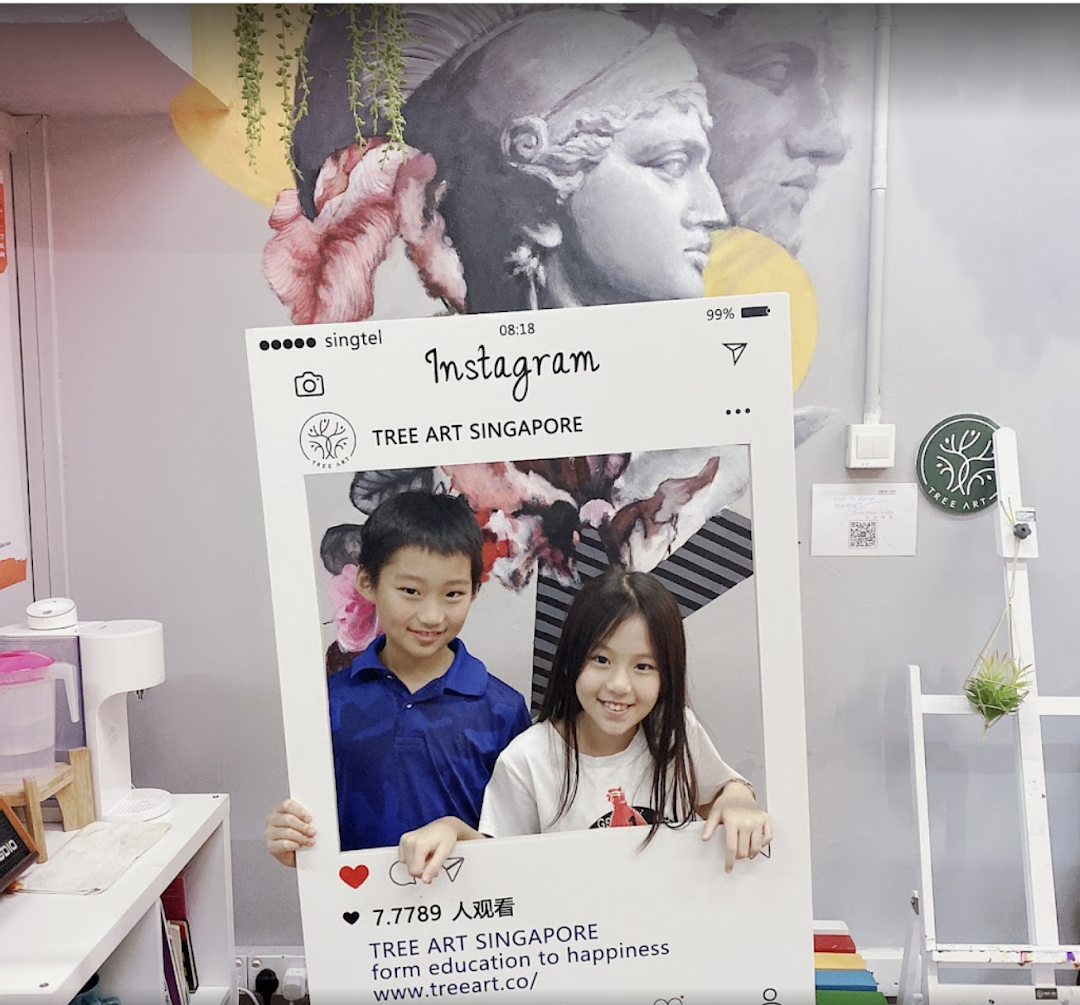 Two smiling children pose with an Instagram frame inside the vibrant Tree Art studio, featuring a classical mural.
