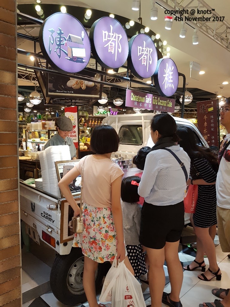 A bustling food stall named TuTu Coconut Cake with customers and staff, featuring a unique truck display.