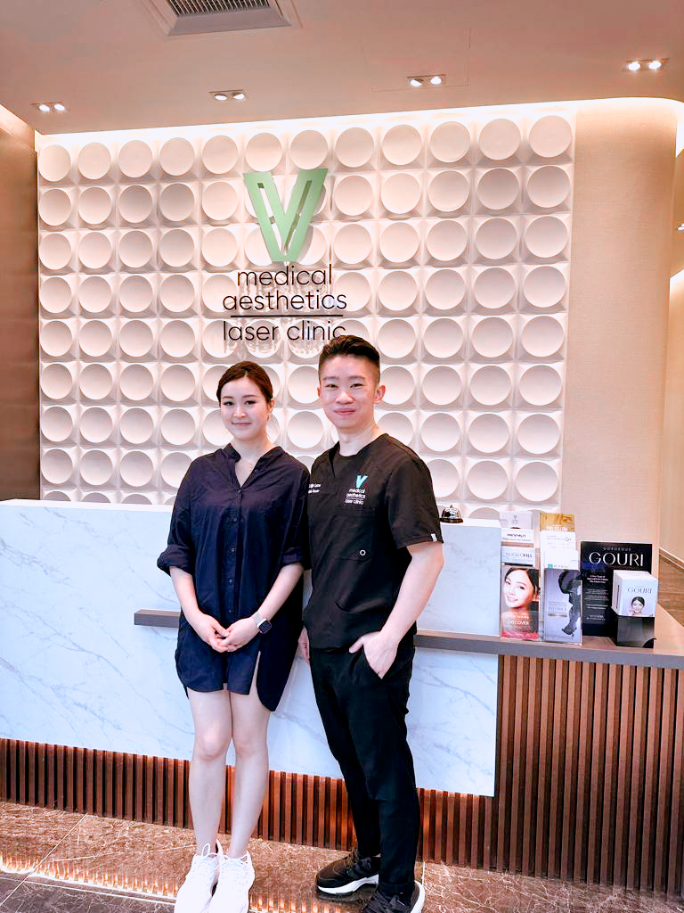 Two individuals stand at the reception of V Medical Aesthetics Laser Clinic, featuring its logo and modern decor.