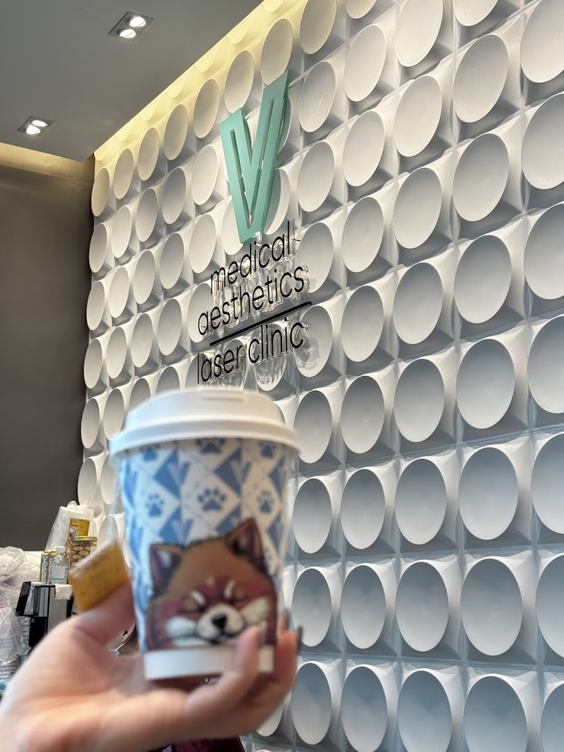 A hand holds a coffee cup in front of a modern textured wall featuring the V Medical Aesthetics & Laser Clinic logo.