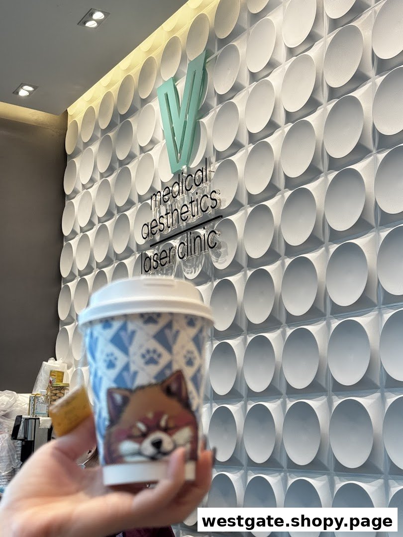 A hand holds a coffee cup in front of a modern textured wall featuring the V Medical Aesthetics & Laser Clinic logo.