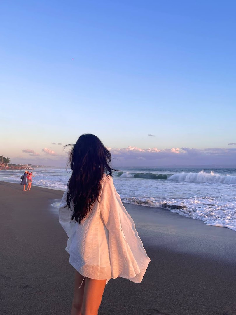 A person with long dark hair stands on a beach, looking at the ocean waves during a beautiful sunset.