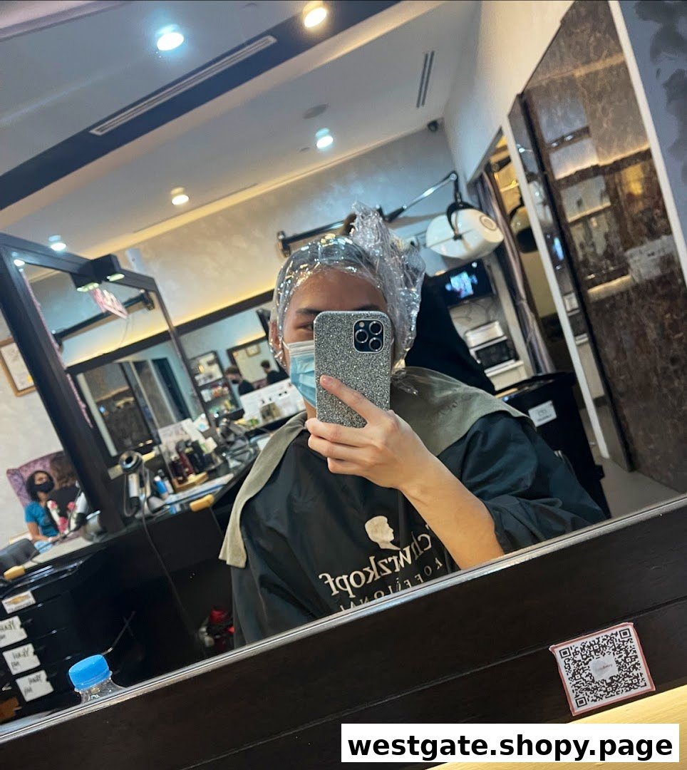 A customer in a salon chair, wearing a hair cap and mask, taking a mirror selfie during a hair treatment.
