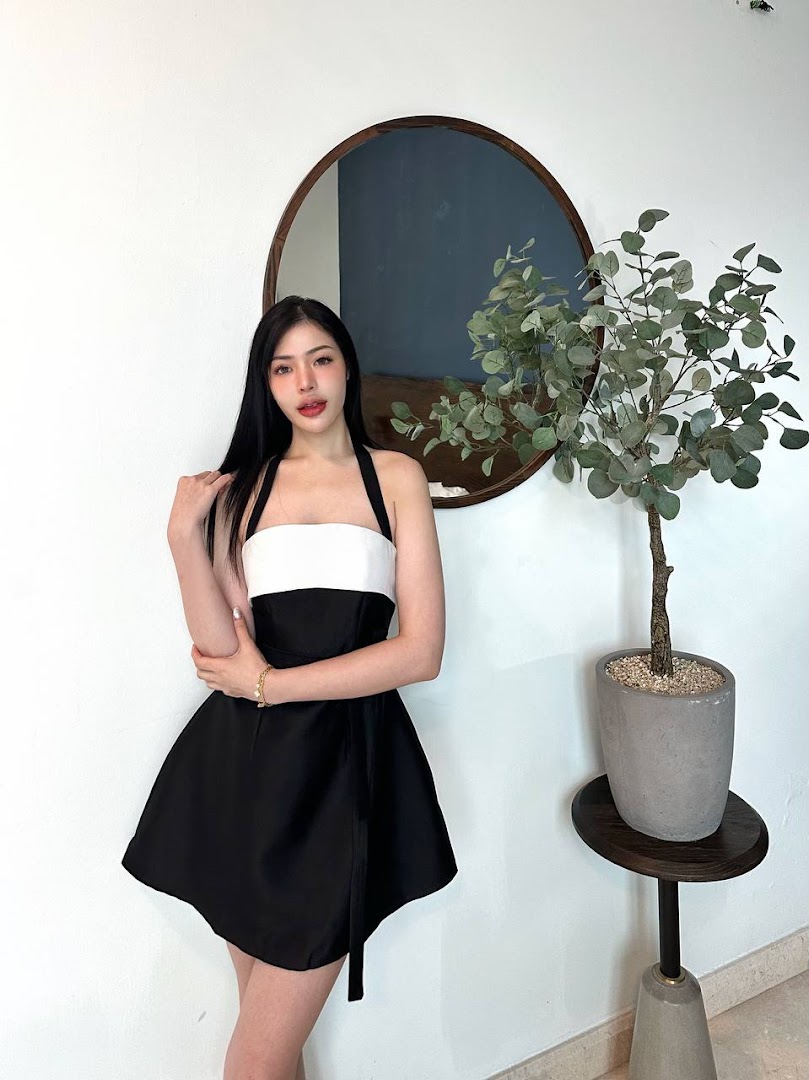 A woman in a black and white dress poses next to a plant and mirror in a stylish interior.