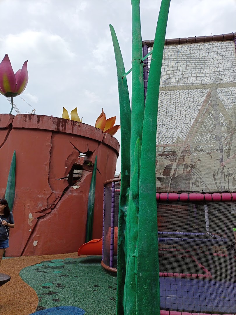 A vibrant outdoor play area featuring giant plant structures, a cracked pot, and a netted climbing frame with a slide.