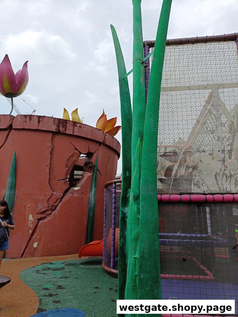 A vibrant outdoor play area featuring giant plant structures, a cracked pot, and a netted climbing frame with a slide.