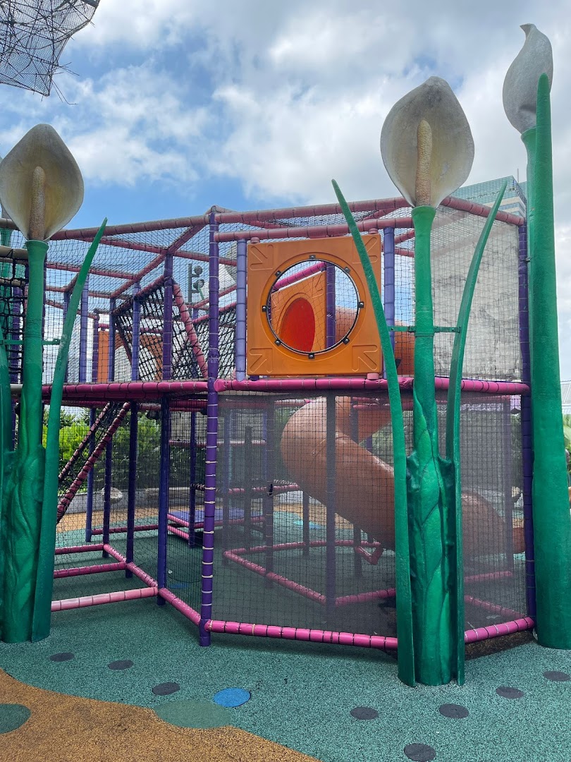 A vibrant outdoor playground with colorful structures, slides, and climbing nets, surrounded by large plant sculptures.