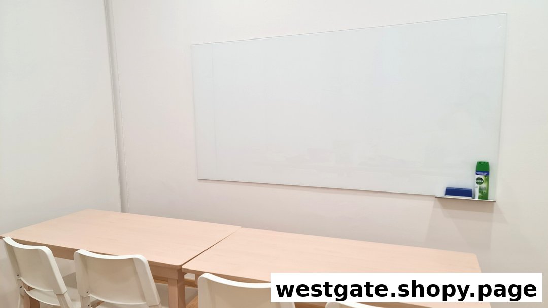 A clean, bright classroom with light wooden tables, white chairs, and a large whiteboard.