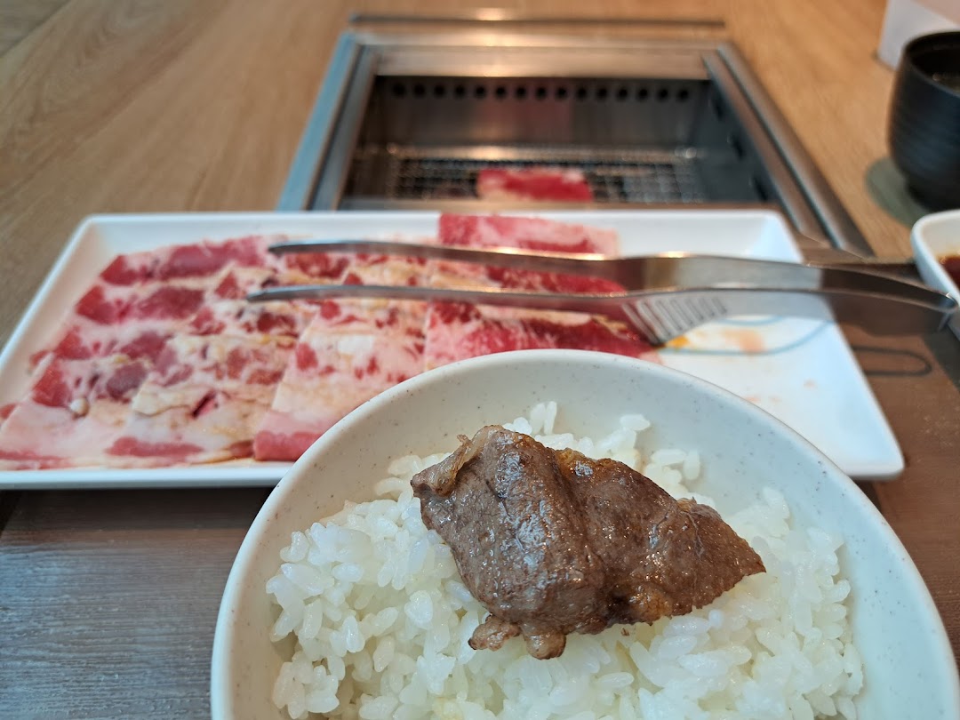 A close-up of a bowl of rice with grilled meat, alongside a plate of raw yakiniku slices and a grill.