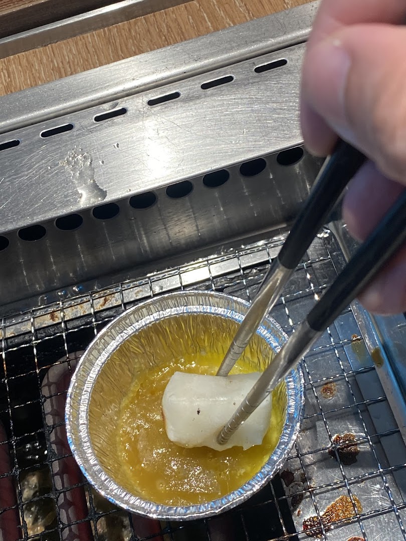 A hand uses tongs to dip a food item into a sauce on a grill at Yakiniku Like.