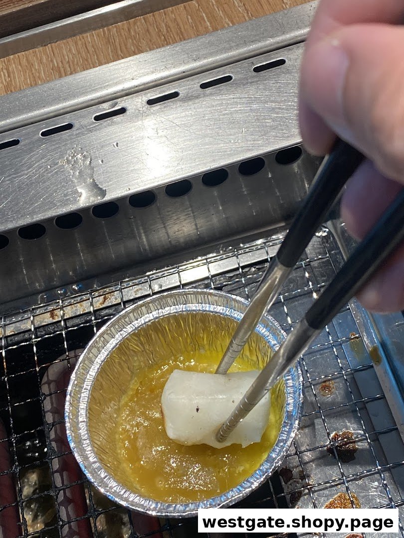 A hand uses tongs to dip a food item into a sauce on a grill at Yakiniku Like.