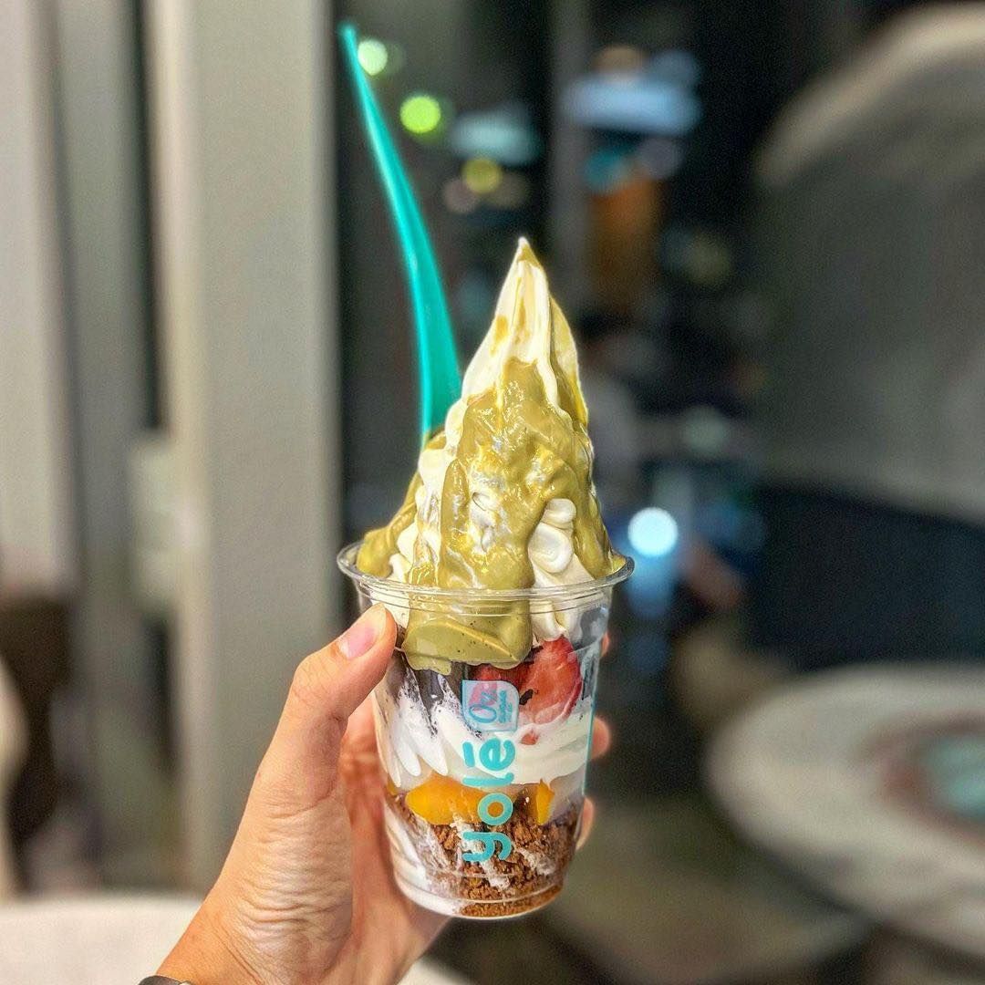 A hand holds a delicious cup of Yolé frozen yogurt with pistachio sauce and fruit toppings.